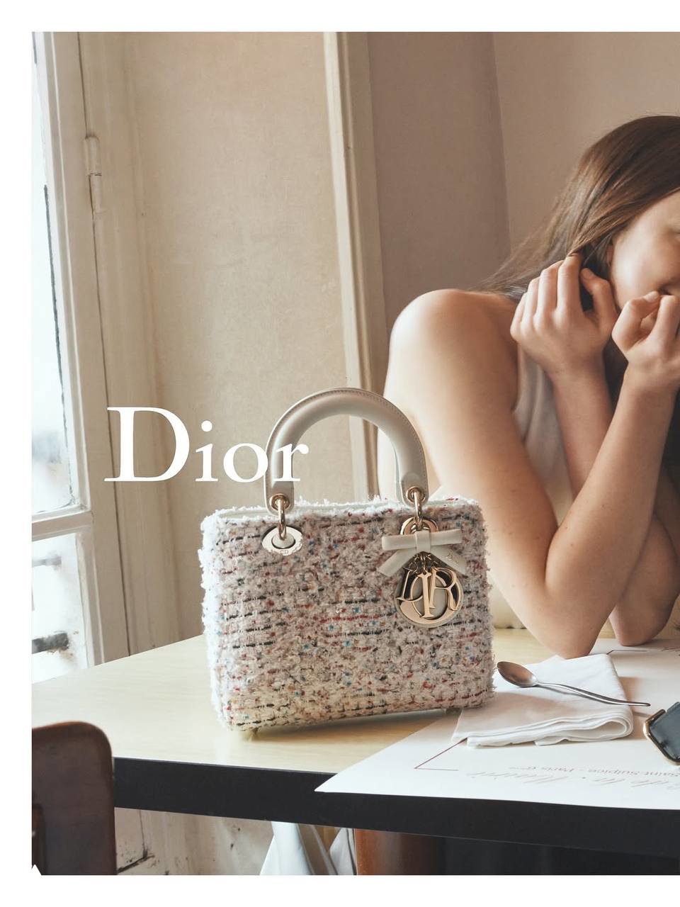 Твидовая сумка Lady Dior на окне кафе, рядом часть силуэта модели — кадр с романтическим тёплым освещением, нежной текстурой твида и металлическими буквами‑деталями на сумке.