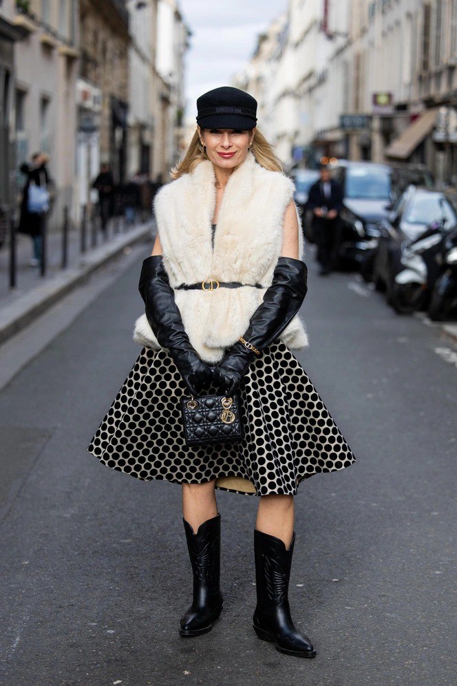Стильная женщина в кремовой пушистой жилетке, зафиксированной ремнём, в юбке с геометрическим принтом и высоких сапогах — фотокадр уличной моды.