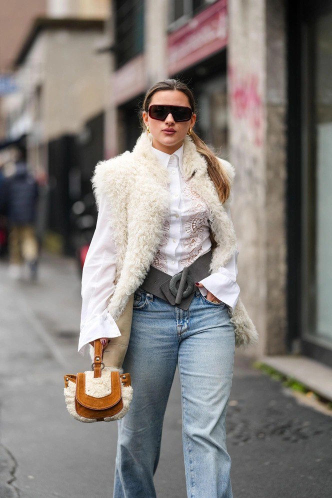 Молодая девушка в белой объёмной меховой жилетке и джинсах, с маленькой сумкой с меховой отделкой — спокойный уличный образ в стиле бохо.