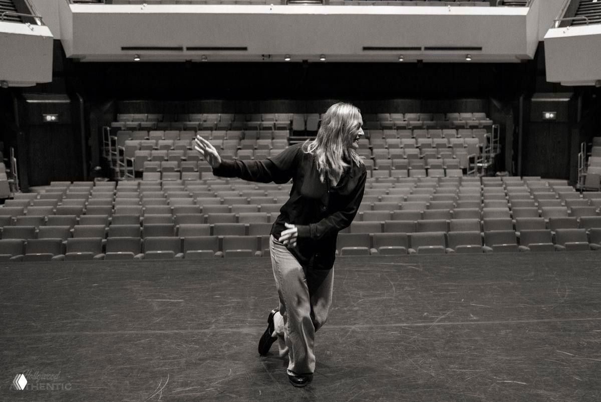Чёрно‑белый снимок на сцене пустого театра: женщина двигается по сцене, вокруг ряды пустых кресел и сцена в полумраке.