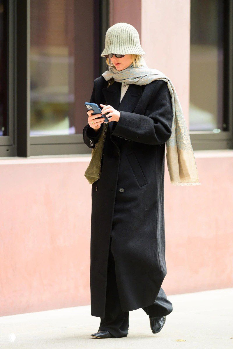 Женщина в длинном чёрном пальто и вязаной светлой шапке, с широким шарфом и солнцезащитными очками, идёт по тротуару и смотрит в телефон.