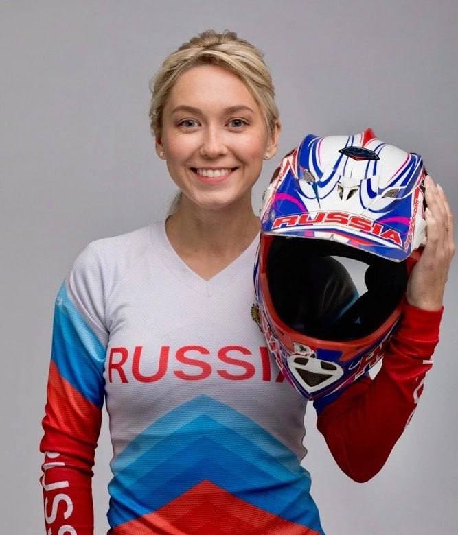 Женщина в спортивной рубашке с элементами флага держит шлем в руке и улыбается, поза демонстрирует спортивный реквизит и неформальную атмосферу.