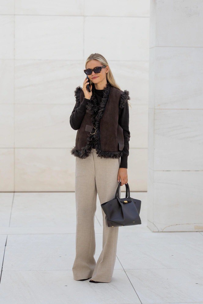 Блондинка в тёмной дублённой жилетке с меховой отделкой, светлые прямые брюки и чёрная сумка на фоне минималистичной архитектуры — элегантный образ.