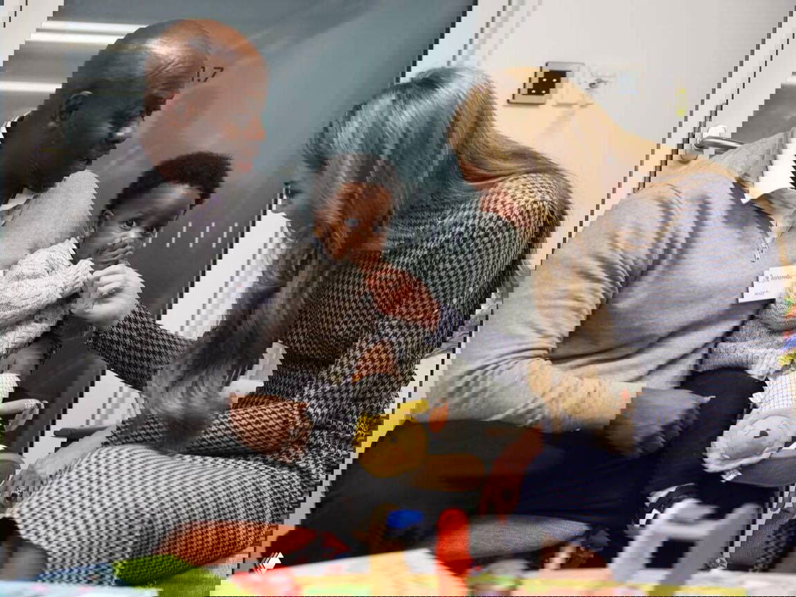 Женщина в ломаной клетке общается с ребёнком в игровой комнате центра поддержки, ребёнок сидит на коленях у мужчины, сценка взаимодействия