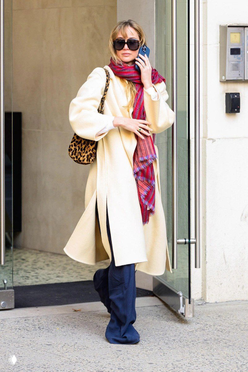 Женщина у входа в здание в светлом пальто, с ярким красным шарфом и леопардовой сумкой на плече; кадр у городского подъезда.