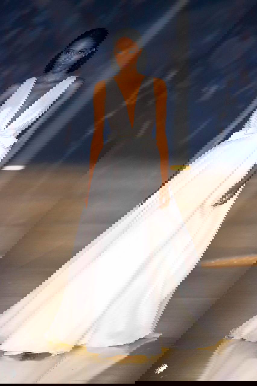 Модель в платье с глубоким вырезом и пышной юбкой, украшенной бантом на талии — классический свадебный силуэт на подиуме.