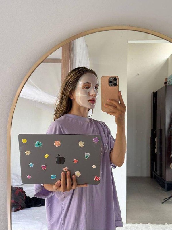 Селфи в зеркале: девушка в тканевой маске для лица держит ноутбук с наклейками — кадр о домашнем уходе и восстановлении кожи лица.