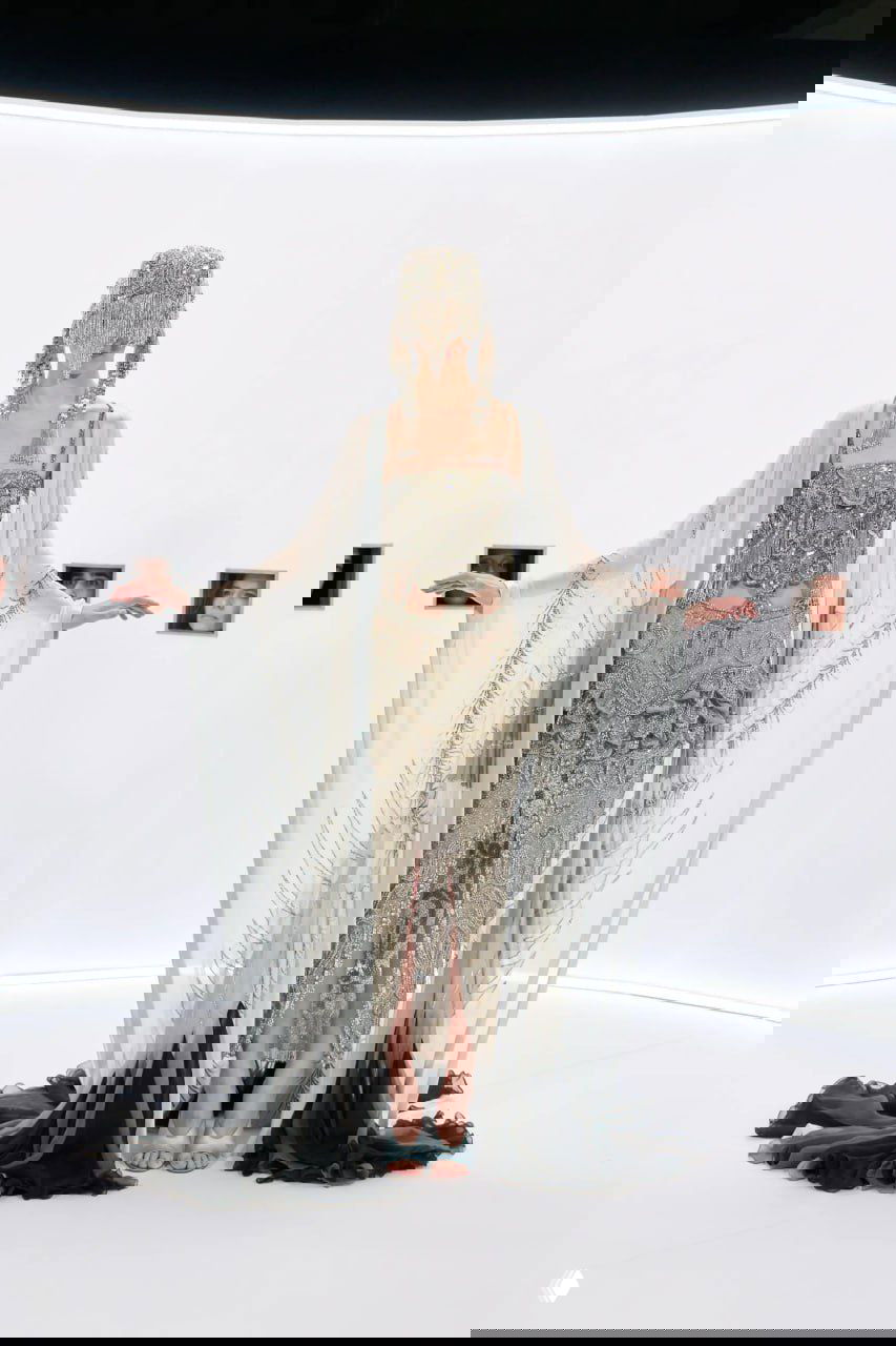 Высокая модель в ярко-розовом платье с фактурной накидкой из перьев на плечах на фоне белой инсталляции показа Valentino.