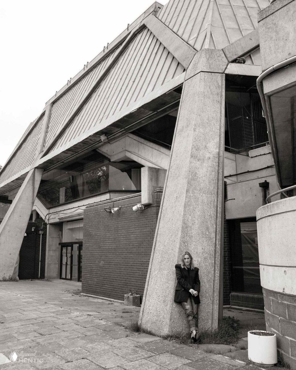 Широкий план у бетонного фасада: фигура актрисы у массивной опоры здания, кадр подчёркивает масштаб и текстуру материала.