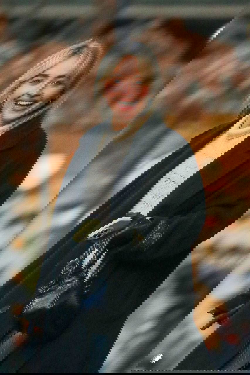 Крупный план женщины в светлом вязаном свитере под чёрным пальто: она держит сумку и телефон на фоне городской витрины.