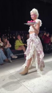 Модель в асимметричном наряде идёт по подиуму, держит поднос с десертами; образ подчёркивает сочетание роскоши и панк-эстетики.