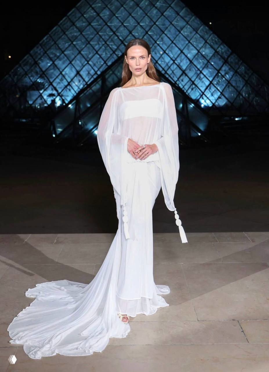 Женщина в легком белом ансамбле с длинным струящимся шлейфом стоит на фоне стеклянной пирамиды Лувра, акцент на прозрачности ткани