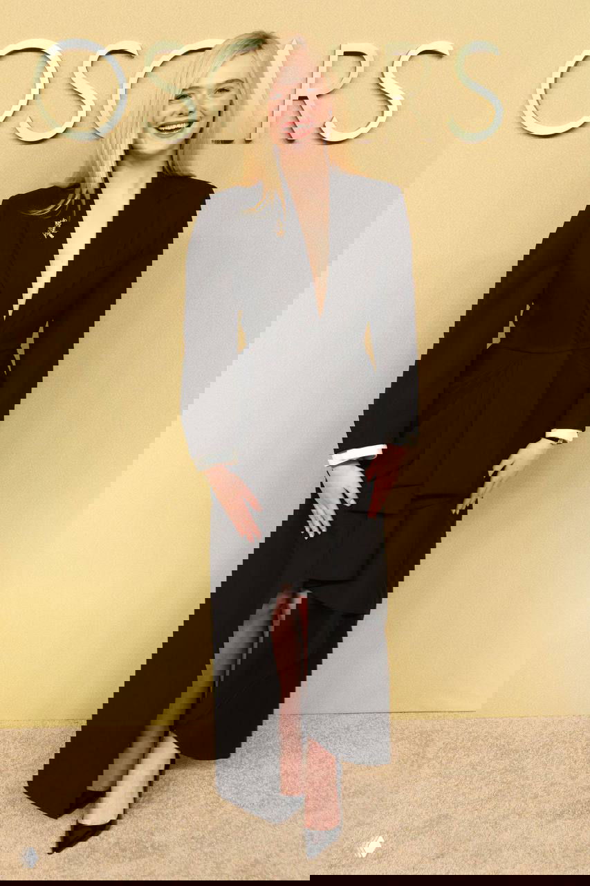 Женщина в длинном чёрном платье с глубоким декольте улыбается на бежевом фоне с надписью OSCARS, вечерний образ на ланче.