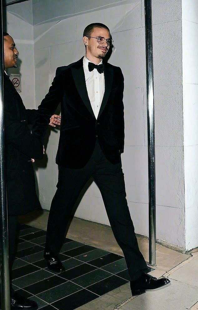 Мужчина в бархатном смокинге и круглых очках выходит из дверей на вечерней прогулке рядом с охраной после афтерпати.