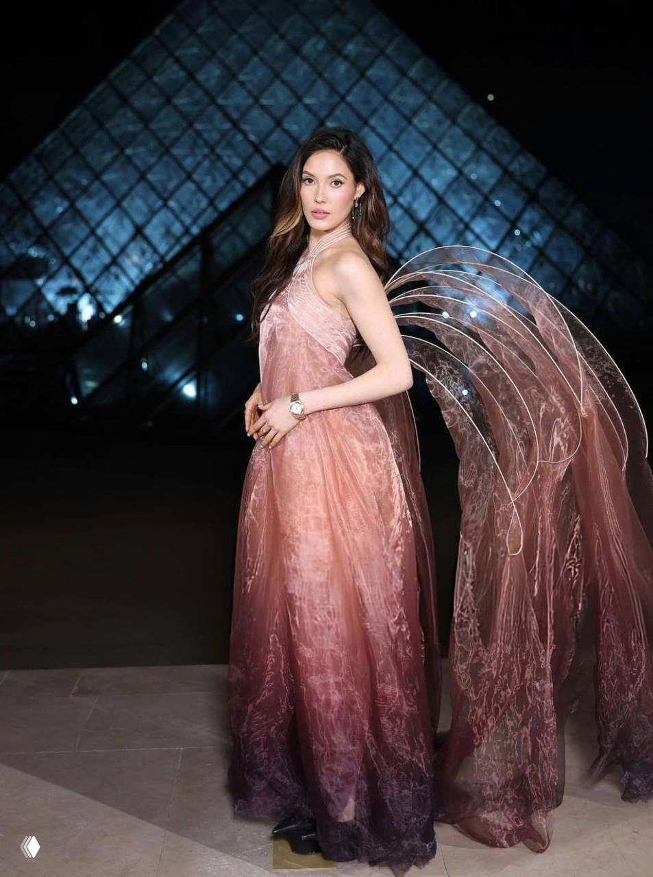 Женщина в розовато-фиолетовом платье с полупрозрачными крыльями-шлейфом позирует перед подсвеченной стеклянной пирамидой Лувра вечером