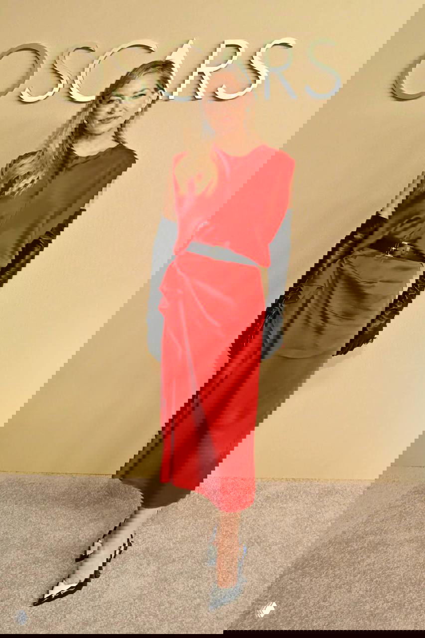 Женщина в красном платье миди с чёрными длинными перчатками позирует у бежевой стены с логотипом OSCARS во время мероприятия.