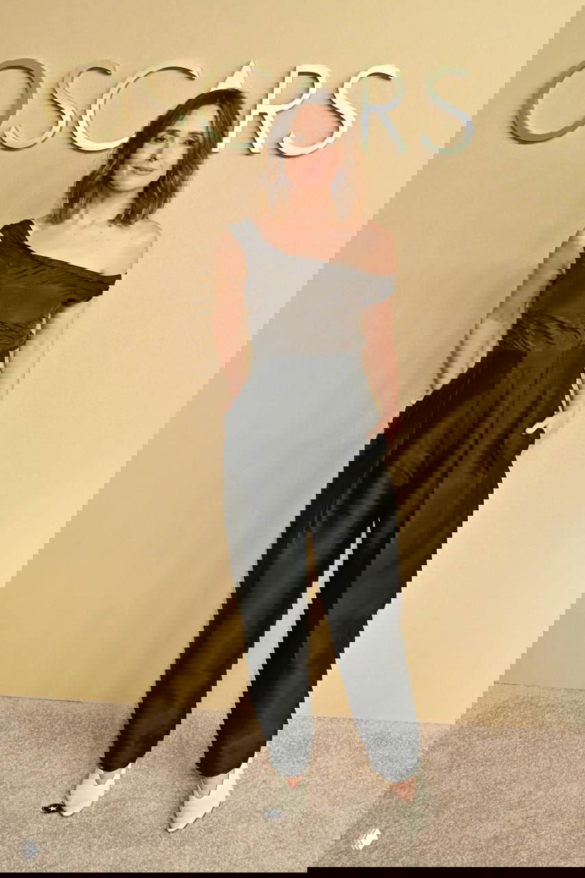 Женщина в чёрном наряде с асимметричным верхом и брюками позирует у стены с надписью OSCARS на ковровой дорожке мероприятия.