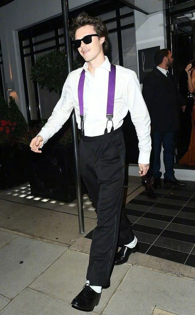 Молодой мужчина в белой рубашке, чёрных брюках и пурпурных подтяжках идёт по тротуару рядом с входом в заведение после вечернего события.