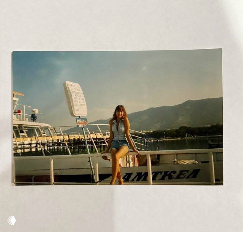 Фото на причале: молодая Ирина Шейк сидит на перилах прогулочного катера на фоне гор и морского пейзажа в солнечный день.