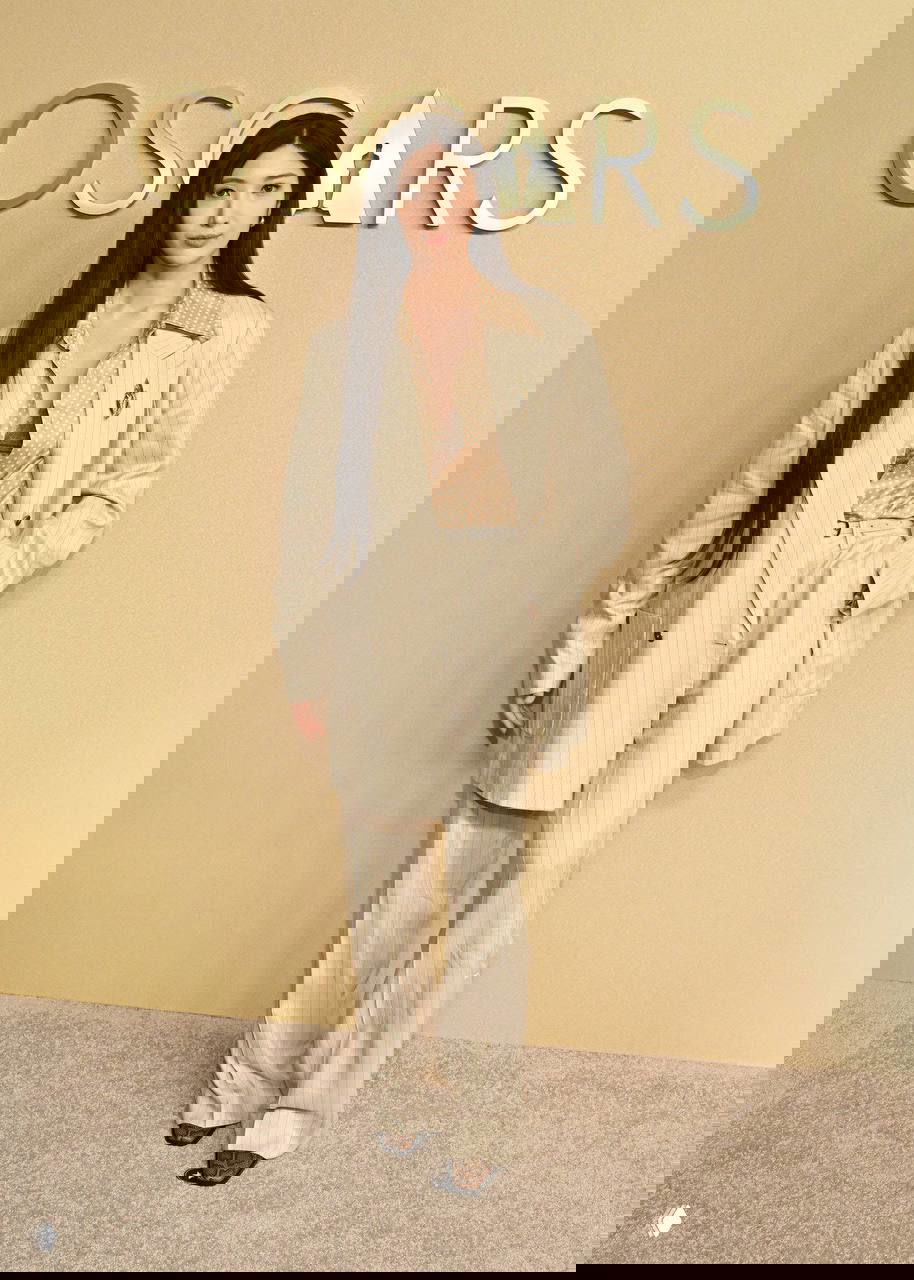 Женщина в светлом брючном костюме с длинными тёмными волосами позирует на бежевом фоне с логотипом OSCARS на мероприятии.