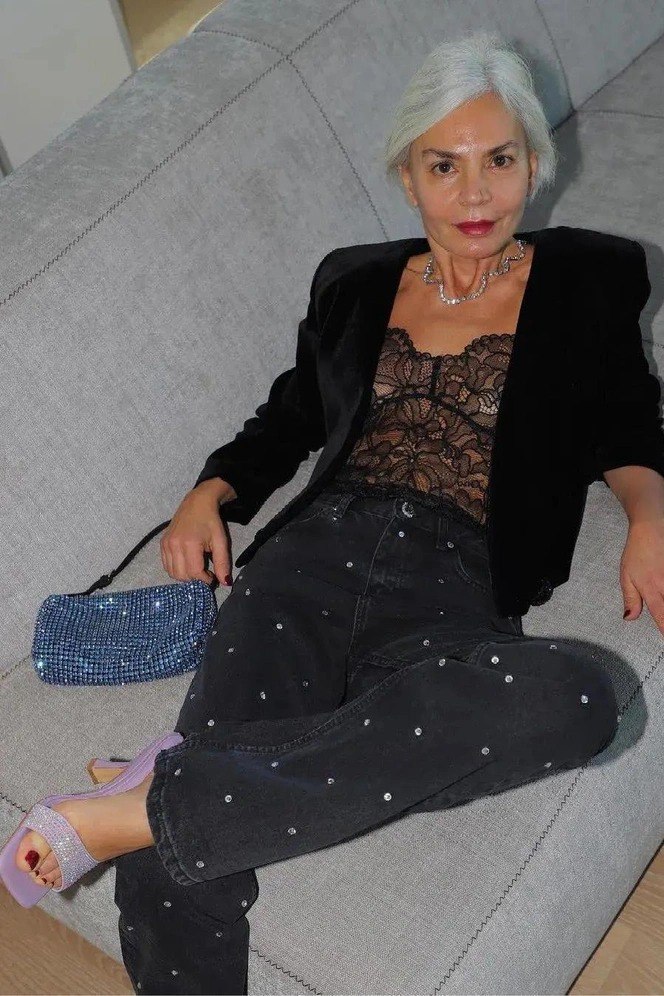 Пожилая женщина с седыми волосами в кружевном топе, коротком пиджаке и черных джинсах с блёстками отдыхает на диване рядом с маленькой сияющей сумочкой, кадр стилизован под модную съёмку.