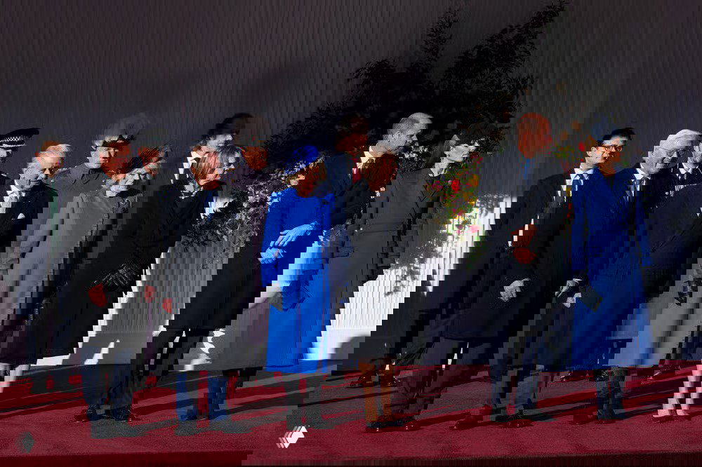 Гости собрались на церемонию у замка: несколько человек в тёмных и синих пальто выстроены на красной дорожке в торжественной обстановке.