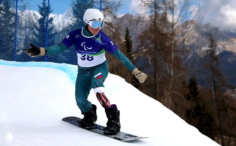 Паралимпийский сноубордист на трассе, в защитном шлеме и в стартовом номере выполняет манёвр на снежном склоне во время соревнований.