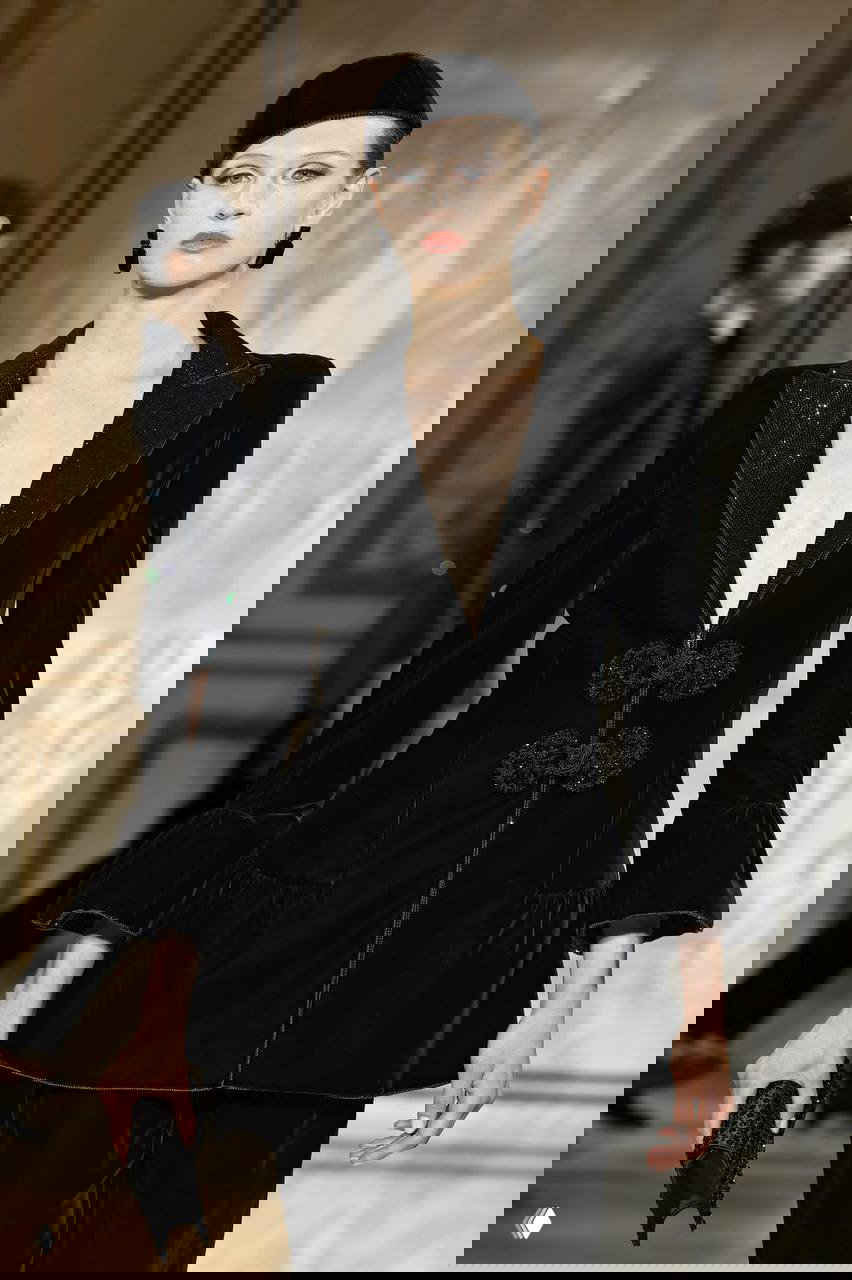 Подиумный кадр: модель в чёрном бархатном жакете с плетёными застёжками-панькоу, сдержанный кутюрный образ и акцент на деталях.