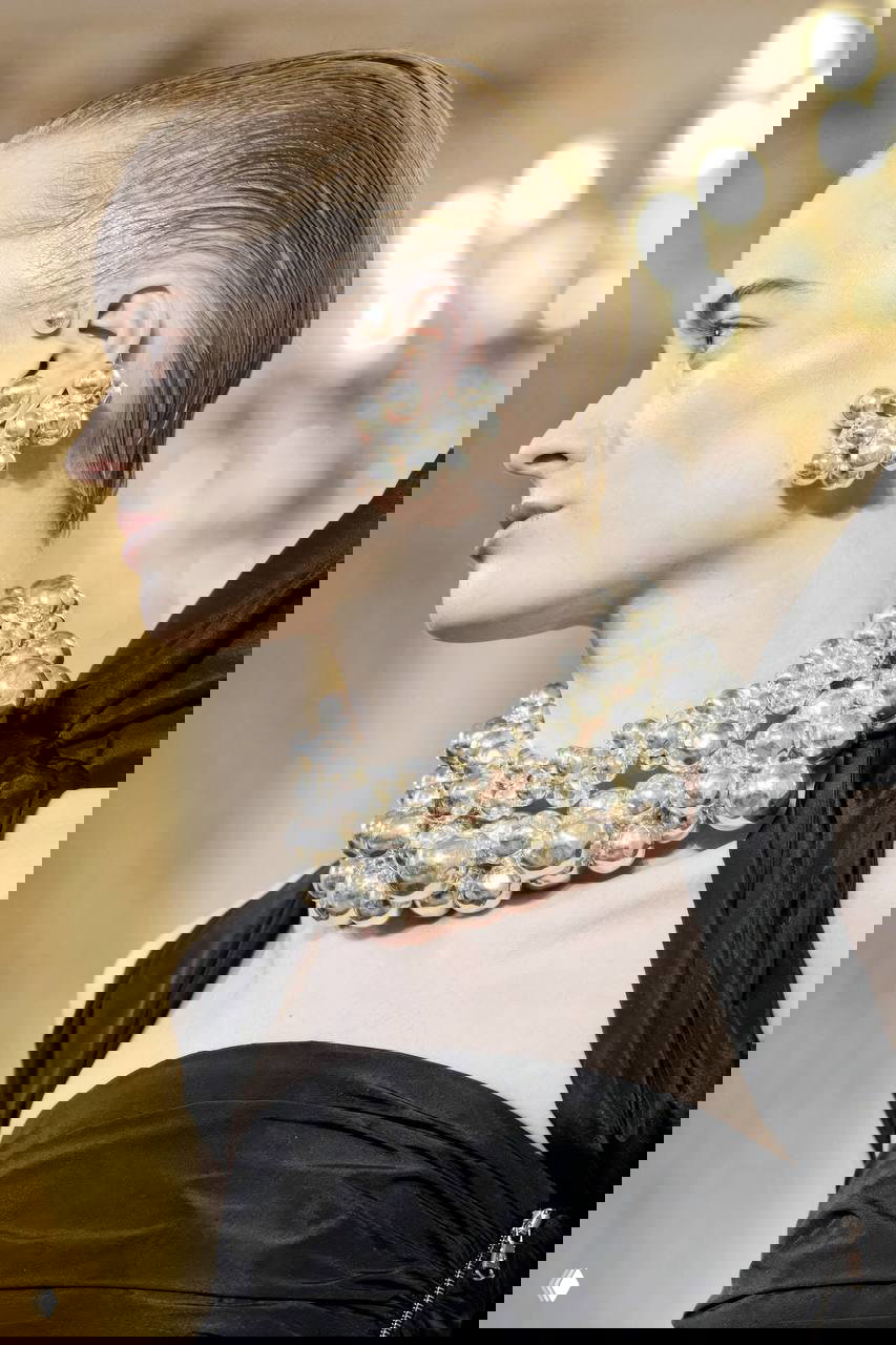 Профиль модели на подиуме: многослойное круглое жемчужное колье и крупные серьги, фон — зал с люстрой, акцент на блеске.