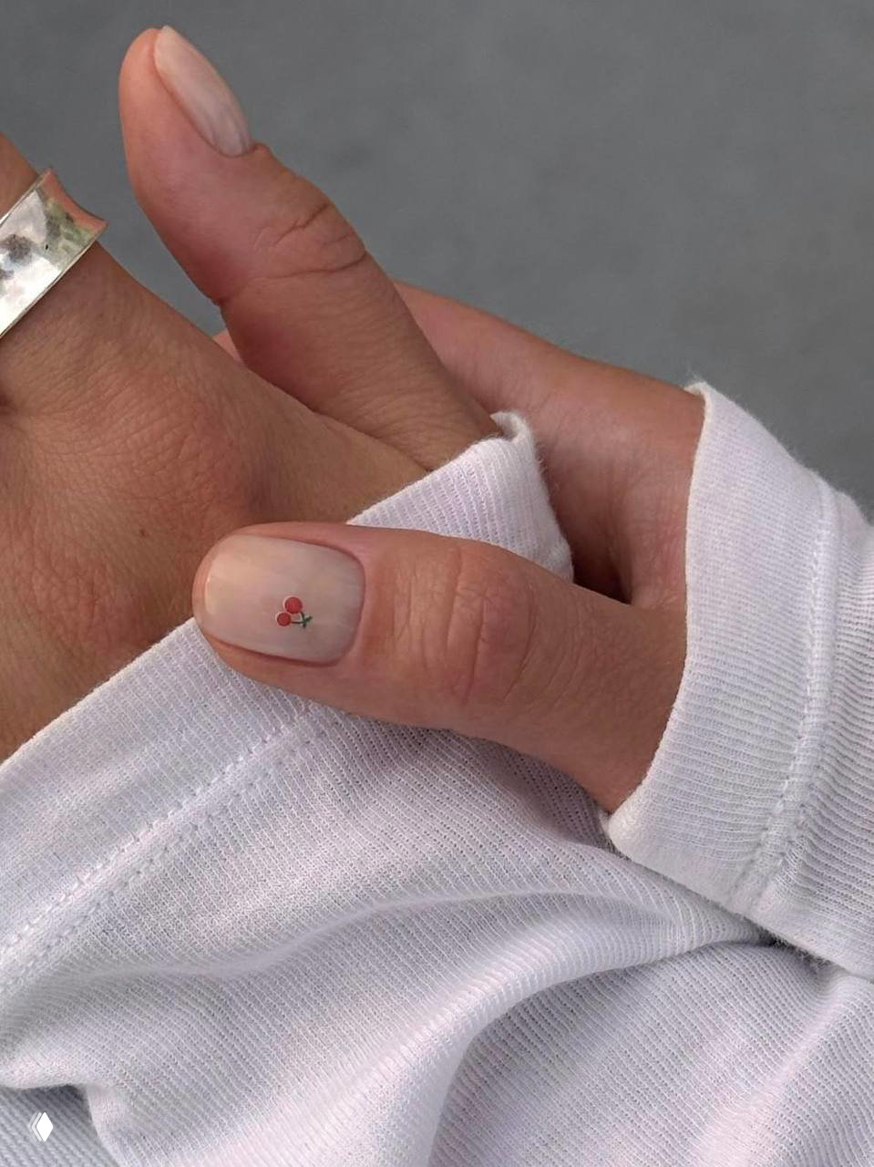 Крупный план большого пальца с нюдовым лаком и миниатюрным дизайном — маленькая вишенка на ногте, белая ткань свитера на фоне.