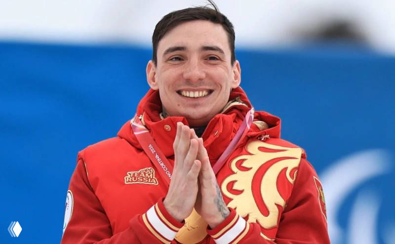 Спортсмен в красной олимпийской форме улыбается вблизи, держит медаль и выглядит растроганным после успешного выступления на Паралимпиаде.