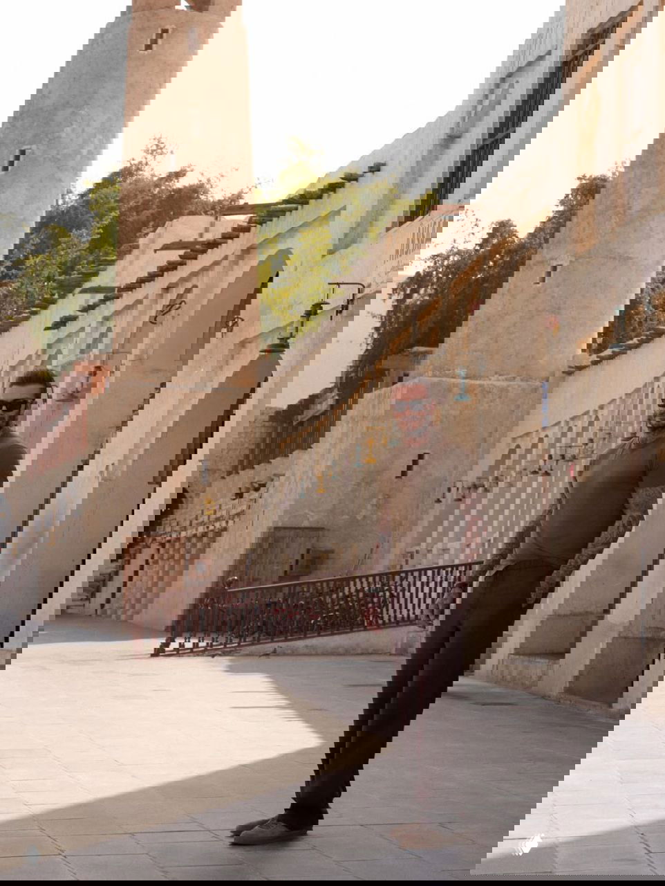 Мужчина стоит у старинной башни и аркадного ряда зданий на пустынной солнечной площади, лёгкая тень от колонн.