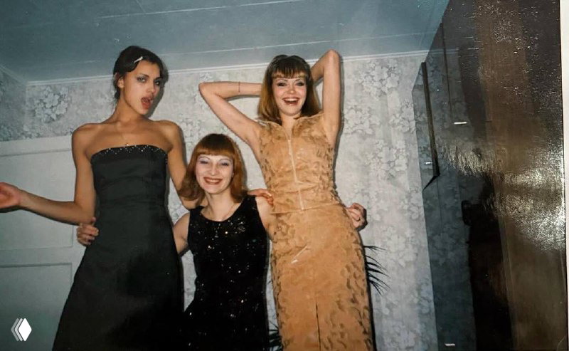 Три подруги на домашней вечеринке: Ирина Шейк и две знакомые улыбаются, нарядные платья и интерьер с цветочными обоями.