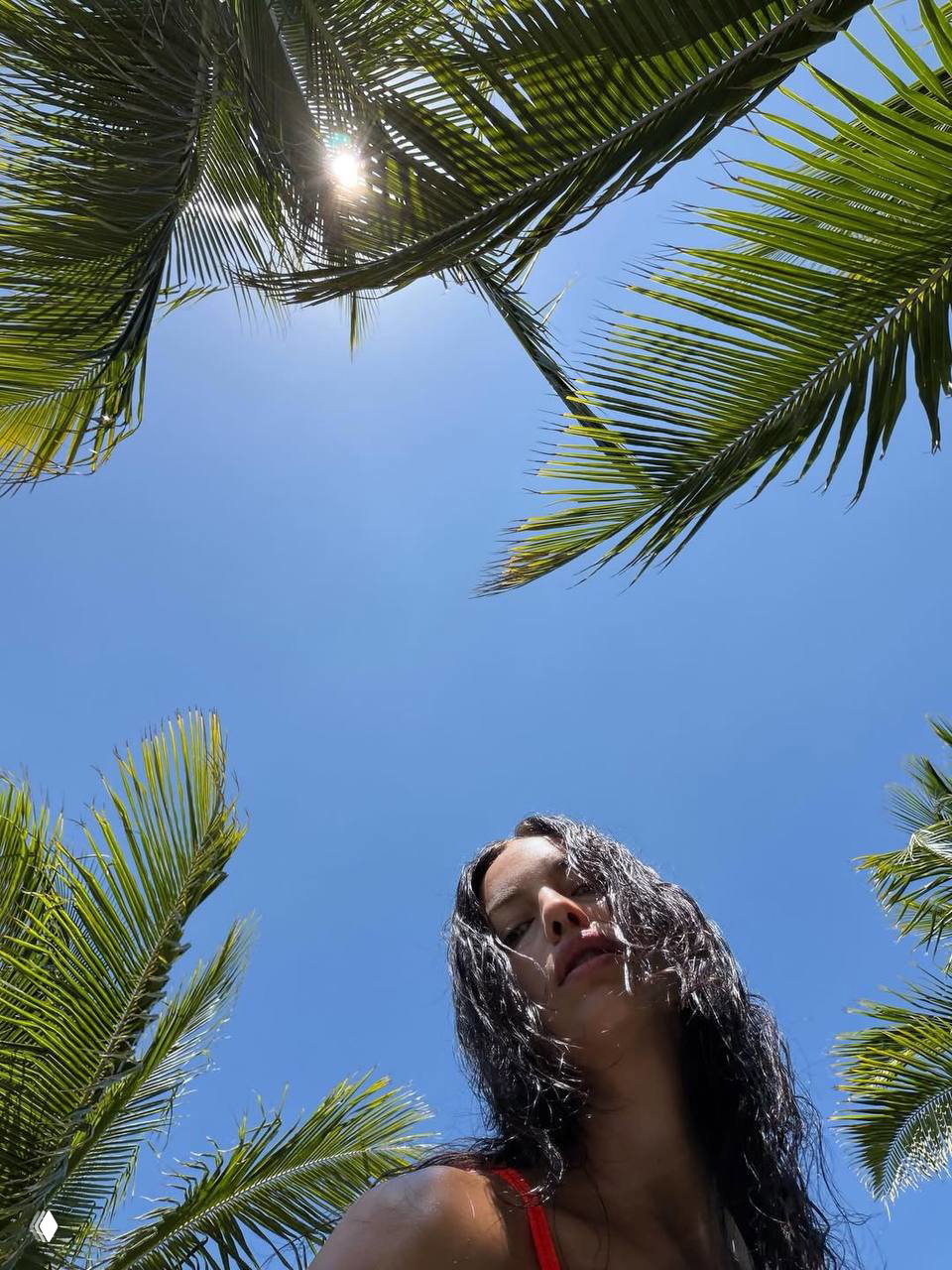 Низкий ракурс: женщина в красном купальнике на фоне голубого неба и пальм, солнечные блики и летняя атмосфера вокруг.