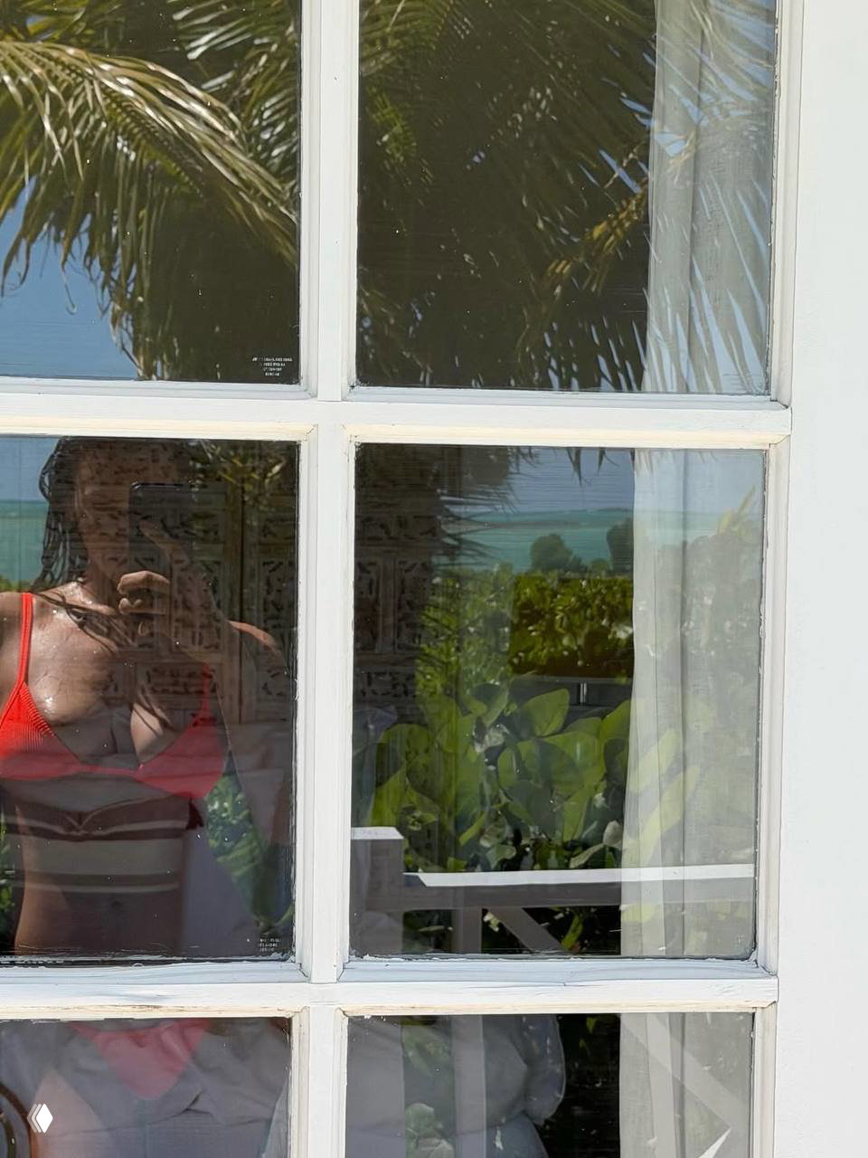 Отражение силуэта женщины в красном купальнике в оконном стекле на фоне зелёных растений и вида на море за окном.