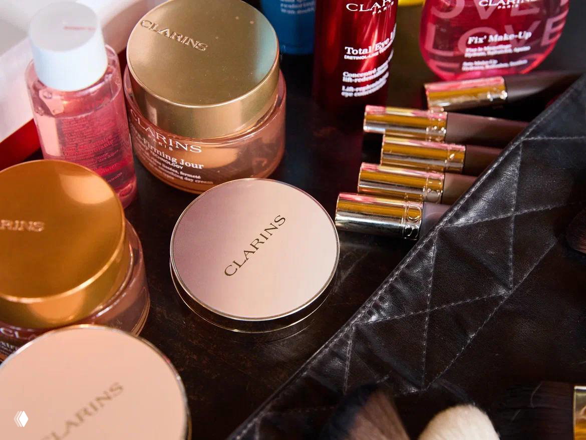 Набор косметики Clarins на столе: баночки кремов, пудры и помады в золотистых и розовых оттенках, вид сверху для демонстрации средств.