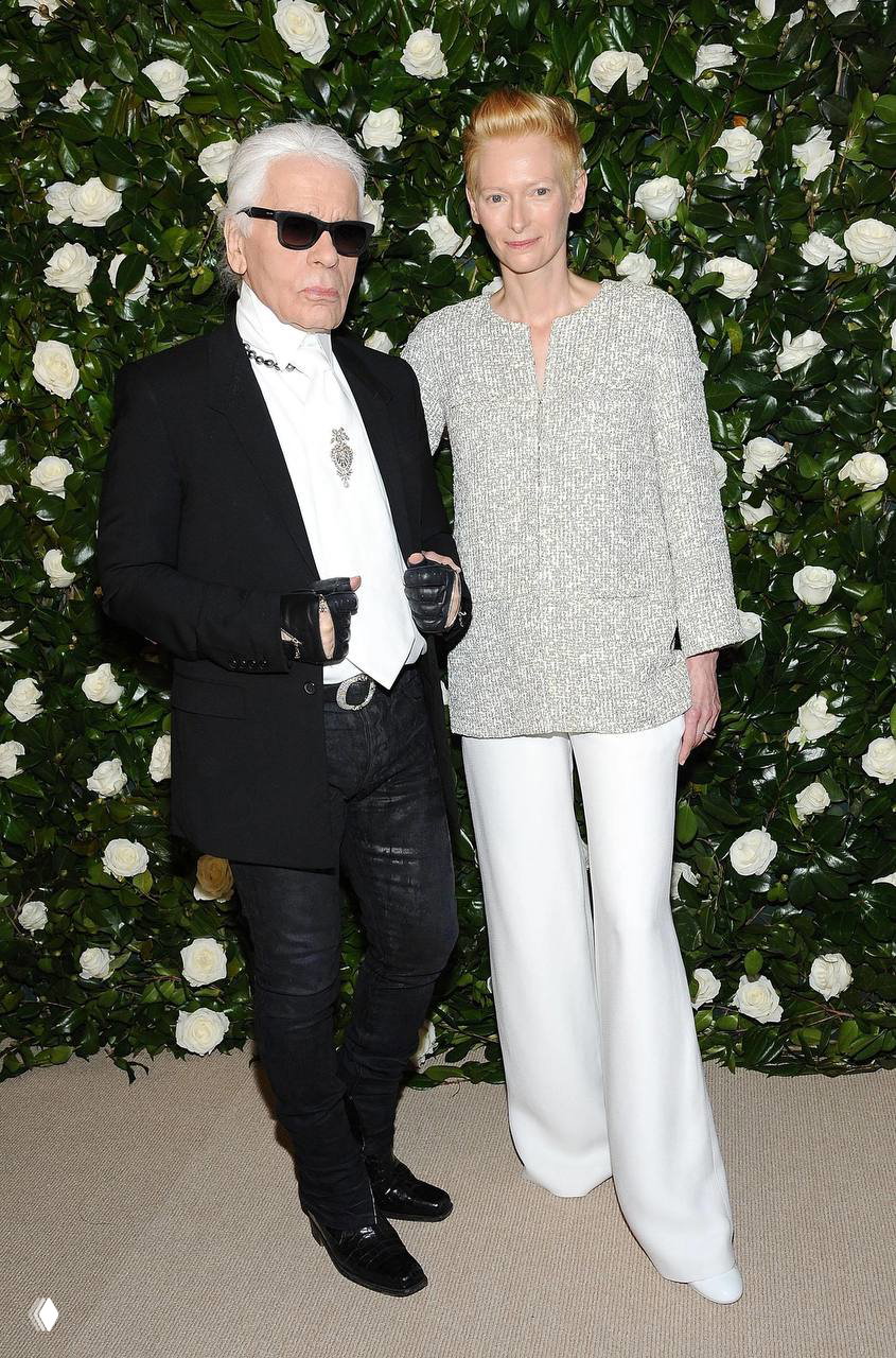 Две фигуры на фоне зелёной стены с белыми розами: женщина в светлом костюме и мужчина в тёмном наряде, фотозона вечернего события и торжественная обстановка.