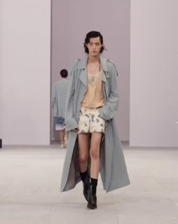 Модель на подиуме в лёгком пальто и шортах, образ показывает игру пропорций и прозрачных топов в мужской коллекции.