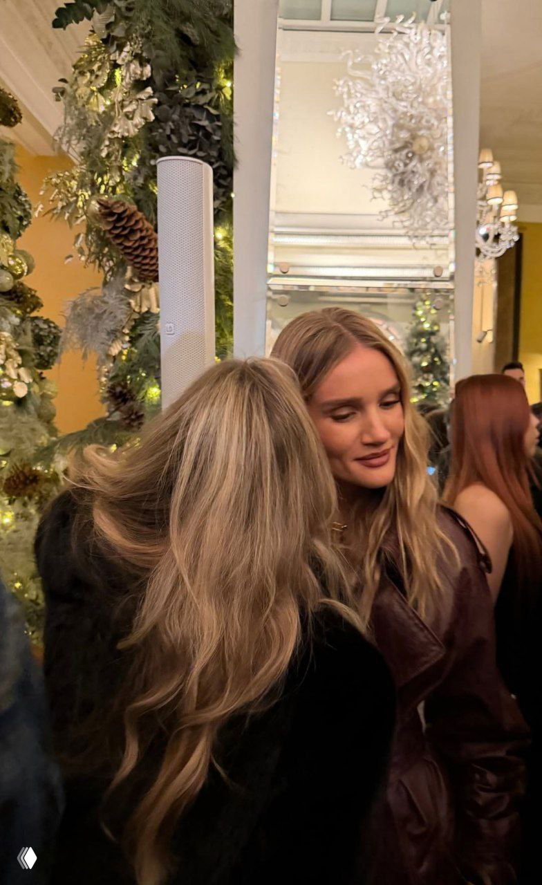 Крупный кадр: женщина в лаковой кожаной верхней одежде общается с гостьей на фоне новогодних декораций и зелёной хвои.