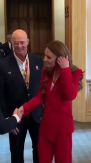 Женщина в алом двубортном костюме дизайнерской марки, приветливо протягивает руку в интерьере с позолотой — приём в Виндзорском замке.