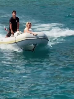 Клава Кока плывёт в надувной лодке по бирюзовой воде: кадр с моря, рядом мужчина на лодке и летняя, солнечная обстановка отдыха.