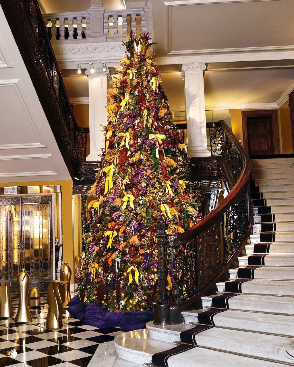 Высокая пёстрая рождественская ёлка, украшенная лентами и сухоцветами, установлена на лестничной площадке гостиничного фойе с чёрно‑белой плиткой.