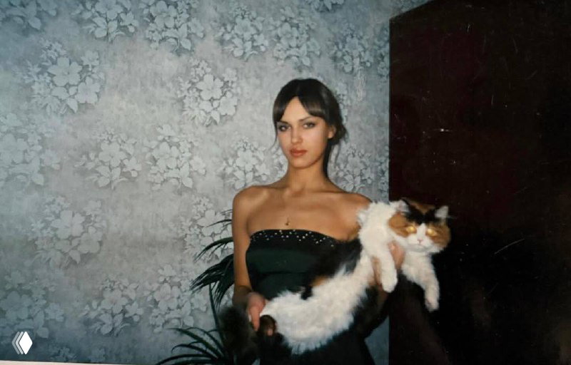 Портрет в интерьере: Ирина Шейк в темном платье держит длинную бело-черную кошку на руках, фон — стена с цветочным рисунком.