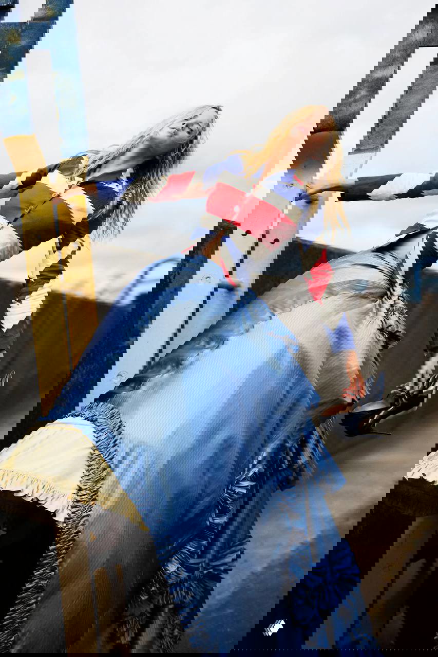 Женщина в жакете с британским флагом и пышной голубой юбкой держит туфли в руке — образ с явными отсылками к эстетике John Galliano и нулевым.