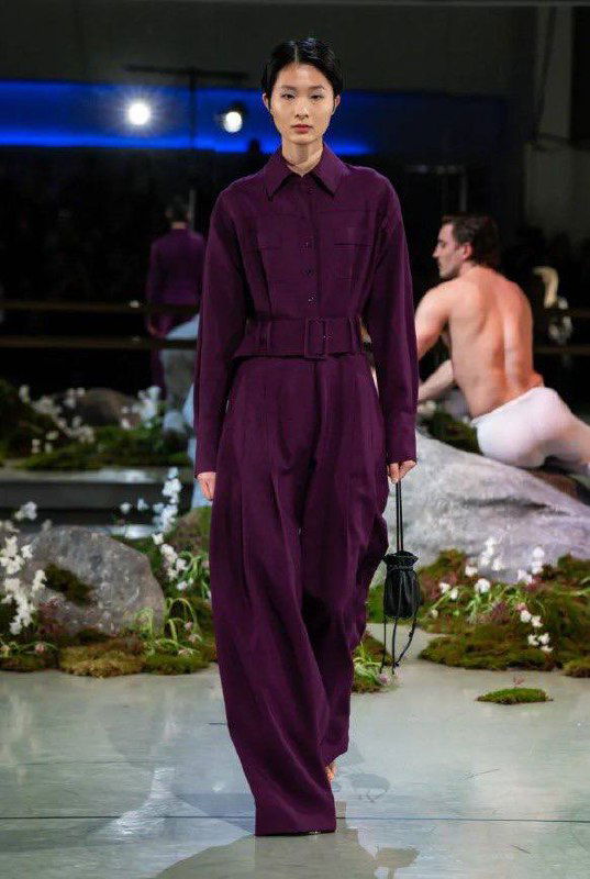 Модель в сливовом брючном комбинезоне с широким поясом шагает по подиуму на шоу Патрика Макдауэлла; строгие линии и глубокий цвет.