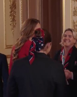 Группа женщин в зале приёма: вид сзади на фигуру в алом костюме и украшенный платок, в окружении гостей и спортивных делегаций.