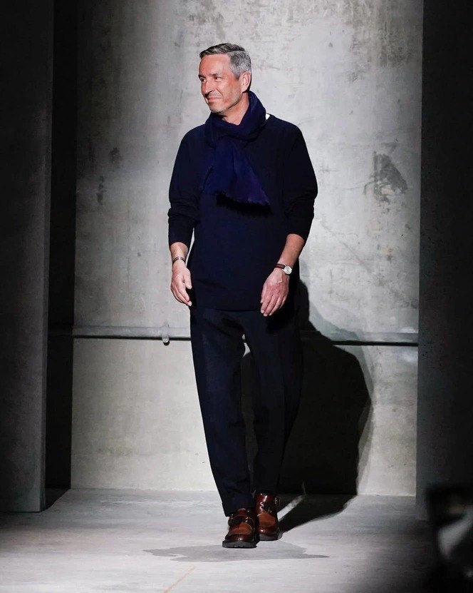 Фронтальный снимок мужчины в темной одежде на подиуме или в студии — полный рост, уверенная поза и деловой образ в нейтральном фоне.