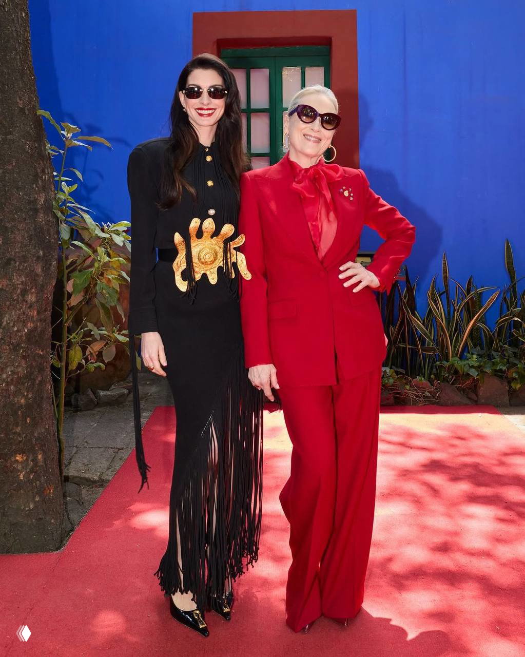Энн Хэтэуэй в чёрном платье Stella McCartney и Мэрил Стрип в ярко‑красном костюме Schiaparelli на красной дорожке в Мехико на фоне синей стены.