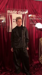 Мужской образ на премии Marie Claire: тёмный костюм свободного кроя на фоне декора с логотипом и бордовыми драпировками сцены.