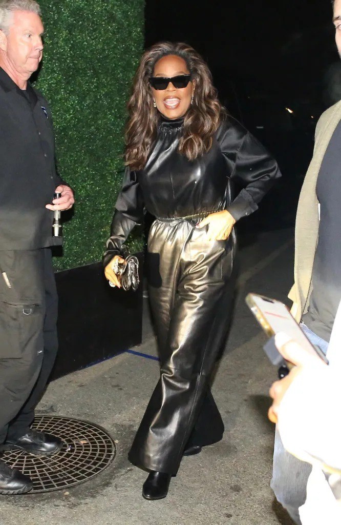 Женщина в чёрном кожаном комплекте с широкими брюками и солнцезащитными очками идёт по улице вечером возле зелёной изгороди.